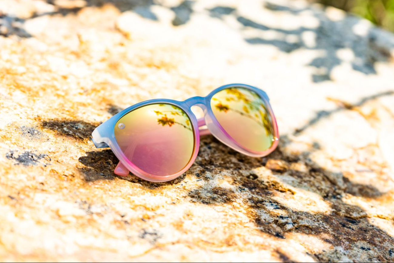 Sunglasses with pink and blue frame and reflective rose gold lenses on a textured rock surface.