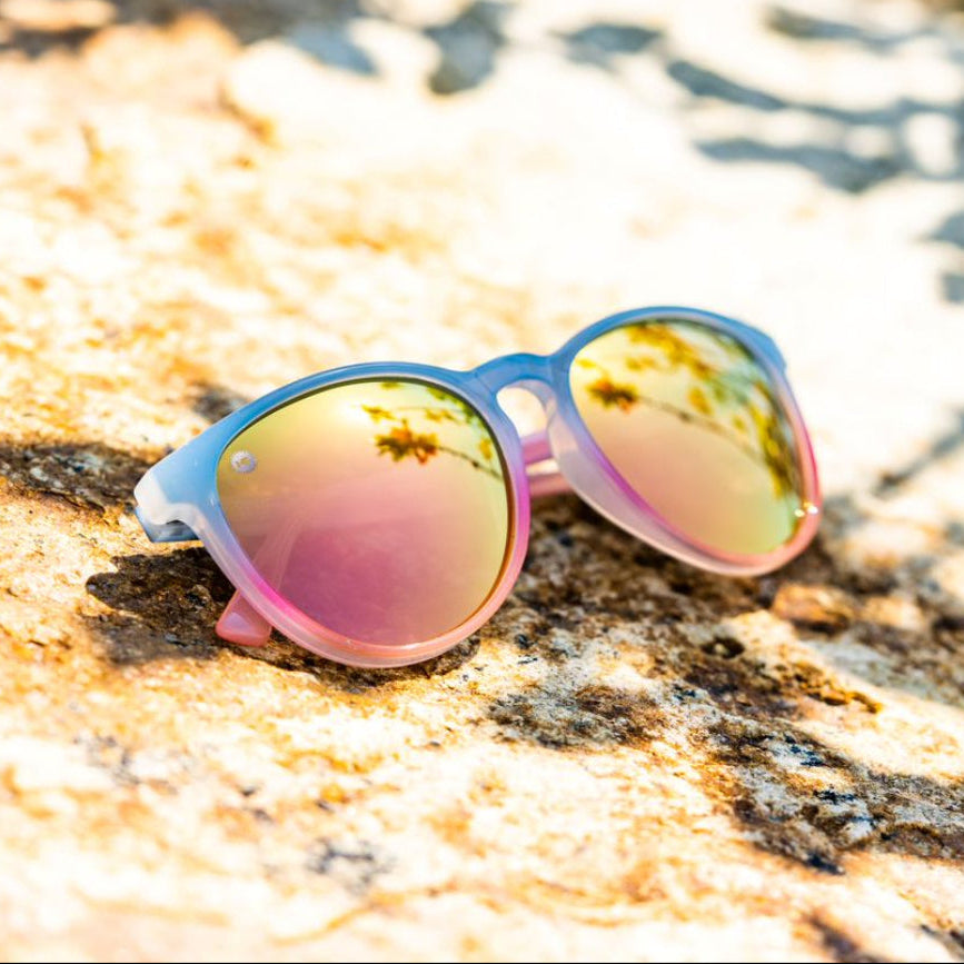 Sunglasses with pink and blue frame and reflective rose gold lenses on a textured rock surface.